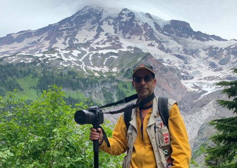 Michael Goodin stands in front of a mountain with a camera